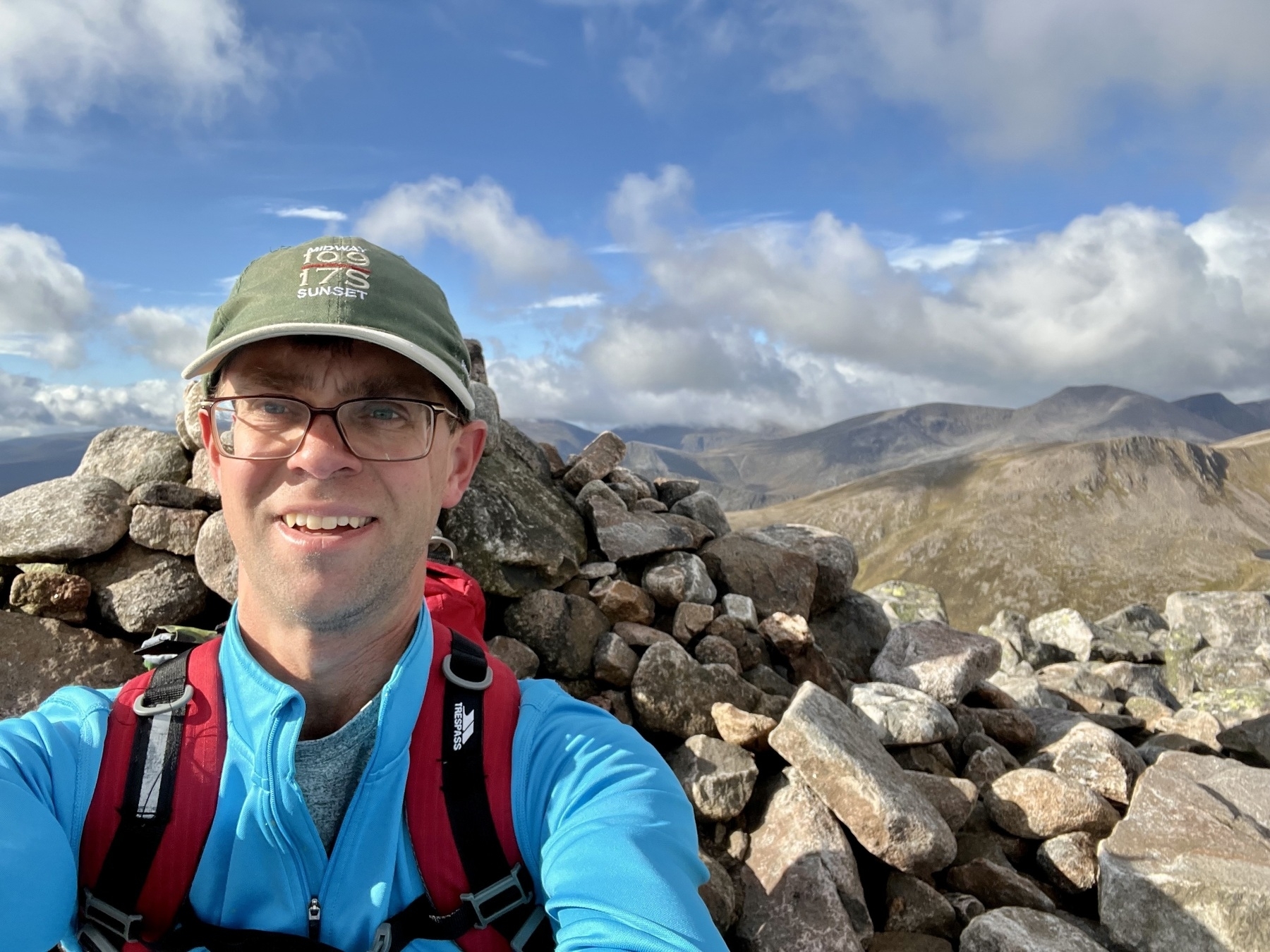 A person wearing hiking gear and a cap is standing on a rocky mountain path with a mountainous landscape in the background.
